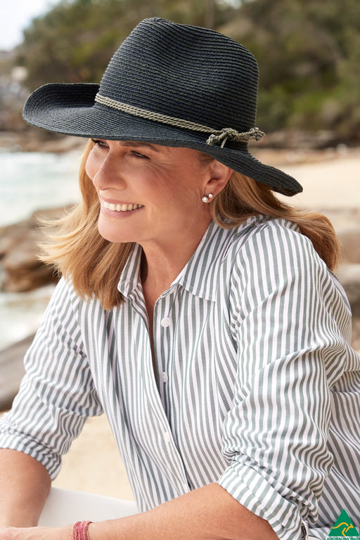 A woman wearing a black woven wide-brim sun hat with a rope detail and a striped button-up shirt, photographed outdoors near a beach or water.