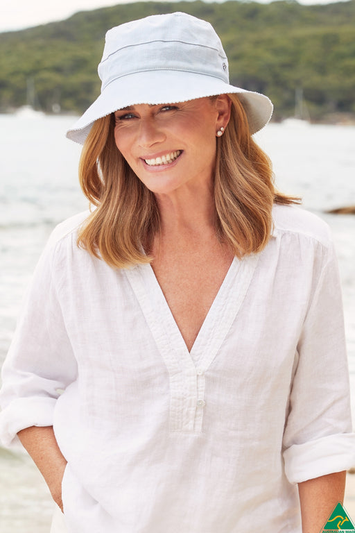 A smiling woman wearing a light blue bucket hat and a white linen shirt stands outdoors near the water with hills in the background.